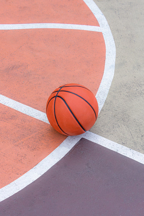 BROOKLYN MESA: A close-up of a basketball resting on the corner of an outdoor basketball court. The ball is orange with black lines and sits on the line of the court.