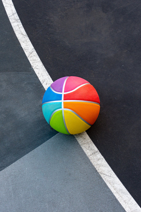 OZ: A rainbow-colored basketball sits on a black and gray court, resting on the edge of a white line. The basketball is brightly colored, with each panel displaying a different shade of the rainbow. The black and gray court is typical of an outdoor basketball court, and the image captures a moment of simple playfulness.