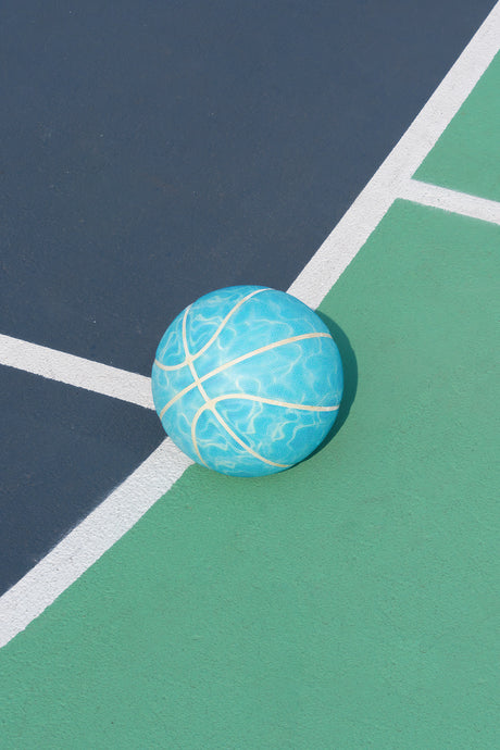 PACIFIC: This image shows a blue basketball resting on a green and black court. The ball is in the corner of the court, with the white lines of the court visible behind it. The ball has a blue and white pattern and appears to be made of rubber.