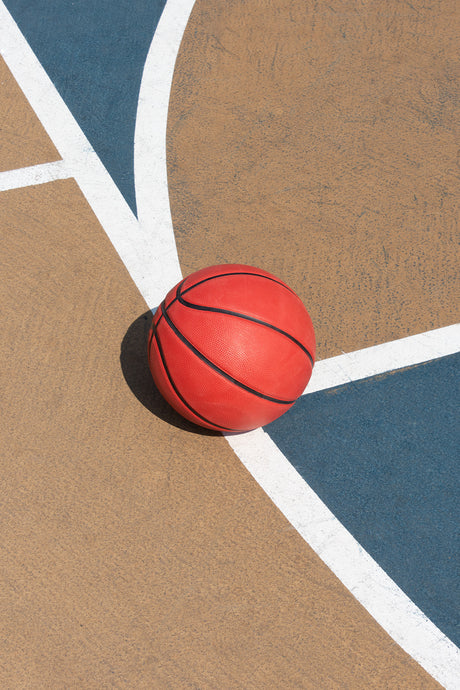 SAND & SEA: A red basketball sits on a basketball court, resting on the corner where two white lines meet. The court is made of a tan material with a blue section in the background.