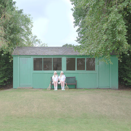 TIME FOR A REMATCH 2: Two women in white tennis dresses sit on a bench outside of a green wooden tennis clubhouse. The women have on white hats and are holding tennis rackets.