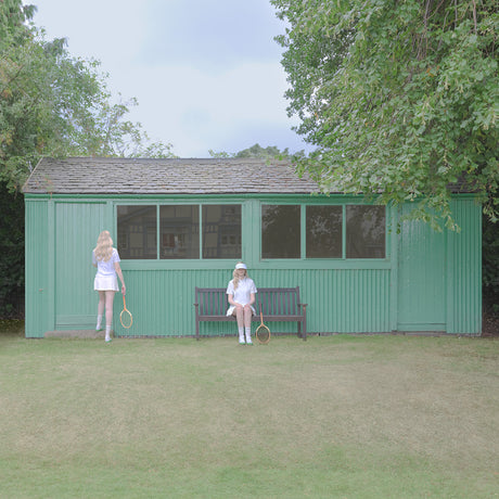 TIME FOR A REMATCH: This image shows two women playing tennis outside a green shed. The woman on the left is holding a tennis racket and is walking towards the shed. The woman on the right is sitting on a bench in front of the shed and is holding a tennis racket. Both women are wearing white tennis outfits. The shed has a wooden roof and is surrounded by green grass.