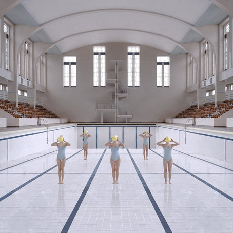 WATER POLO TEAM AT BON ACCORD: A photograph of six women wearing swimsuits and holding yellow balls in front of their faces. They are standing in the shallow end of a large, indoor swimming pool with white tiles and black lane lines. The pool is surrounded by bleachers and a high ceiling with large windows, suggesting a gymnasium or public swimming facility.