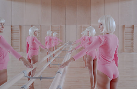 YOU, ME AND THEM: A woman in a pink bodysuit practices ballet at a barre in a studio with large mirrors on the walls. She is facing the camera with her back to the barre, her arms outstretched and her hands touching the barre. She is wearing a white wig and has a focused expression on her face. The studio is brightly lit and the floors are wooden.