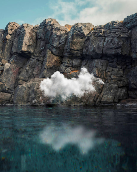 CLOUD II: A large white cloud erupts from the base of a cliff, creating a stunning contrast against the dark rock. The cloud is reflected in the water below, which has a deep blue hue. The sky is clear and blue, with wispy white clouds scattered across it.