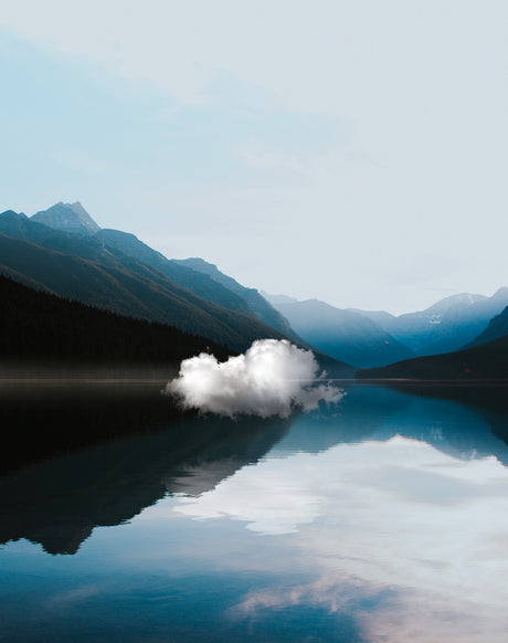 SOMETHING JUST LIKE THIS: A lone, fluffy cloud hovers over a serene mountain lake, its reflection creating a mirror image on the still water. The surrounding mountains are silhouetted against a pale blue sky, adding to the tranquil atmosphere of this picturesque scene.