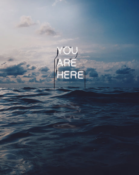 YOU ARE HERE: The image shows a neon sign that reads You Are Here standing in the ocean at sunset. The sky is a mix of blue and grey, with white clouds floating across. The ocean is dark blue and choppy. The sign is a stark white against the dark background, creating a strong visual contrast.