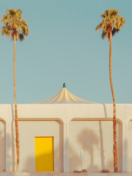 CIRCUS: A photo of a white building with a yellow door flanked by two palm trees. The building has a striped tent-like roof with a bright blue sky in the background.