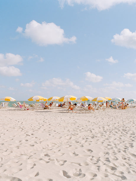 MIAMI BEACH II: A wide shot of a white sandy beach in Miami Beach, Florida. The beach is lined with yellow and white striped umbrellas, under which people are relaxing in beach chairs. The sky is blue with white clouds and the sun is shining brightly.