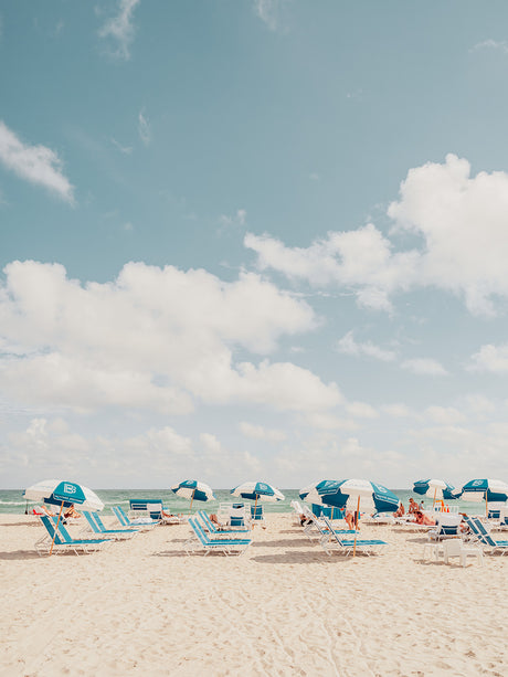 MIAMI BEACH IV: A photograph captures a serene scene of beachgoers relaxing on lounge chairs under blue and white striped umbrellas. The white sand beach stretches out beneath a bright blue sky dotted with fluffy white clouds. The calm ocean water in the background adds to the idyllic atmosphere.