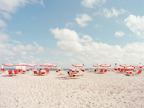 MIAMI BEACH I: The image shows a bright and sunny beach with people relaxing under white and red striped umbrellas. The beach is sandy and the water is clear and blue. The sky is also blue with white clouds.