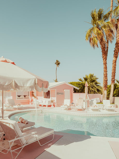 THE PINK OASIS MOTEL: A relaxing poolside scene at a pink motel in Palm Springs, California. The sparkling pool is surrounded by white lounge chairs, palm trees, and a bright blue sky. A pink building with the word Office on it is visible in the background.