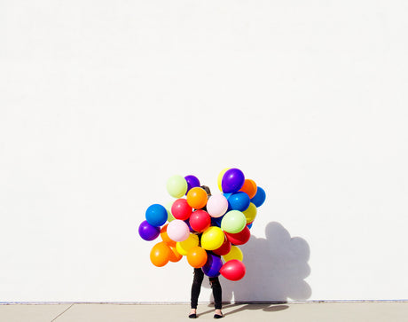 BRIGHT MEDLEY: A person is standing in front of a white wall, holding a large bunch of brightly colored balloons. The balloons are a variety of shapes and sizes, and they are arranged in a loose cluster around the persons body. The persons legs and feet are visible, but the rest of their body is obscured by the balloons. The white wall provides a simple and clean backdrop for the colorful balloons, creating a visually appealing and festive image.