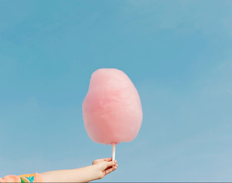 COTTON CANDY SKY: A hand holds a large, pink cotton candy against a clear blue sky. The cotton candy is slightly blurred, but the hand is in focus, emphasizing the sweet treat.