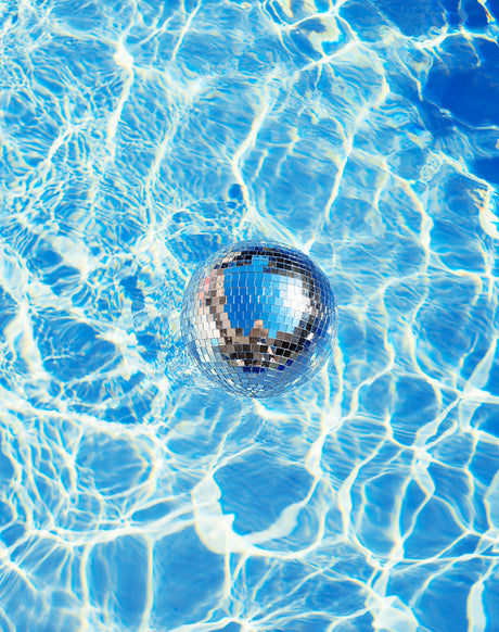 DISCO: A silver disco ball floats on the surface of a blue swimming pool. The water is rippling, creating a pattern of light and dark blue. The disco ball is reflecting the light, creating a dazzling effect.