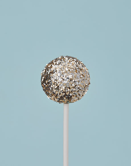 GLITTER DIET LIGHT BLUE: A single cake pop, coated in silver glitter, stands vertically on a white stick against a pale blue background. The cake pop is round and has a smooth, even coating of glitter. The stick is straight and thin, and it extends from the bottom of the cake pop to the bottom of the frame. The background is a solid, light blue color.