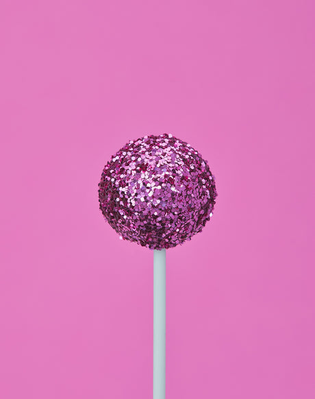 GLITTER DIET PINK: A pink glitter ball on a white stick is set against a plain pink background. The ball has a shiny, sparkly texture, and the stick is thin and straight. The image has a simple and elegant feel.