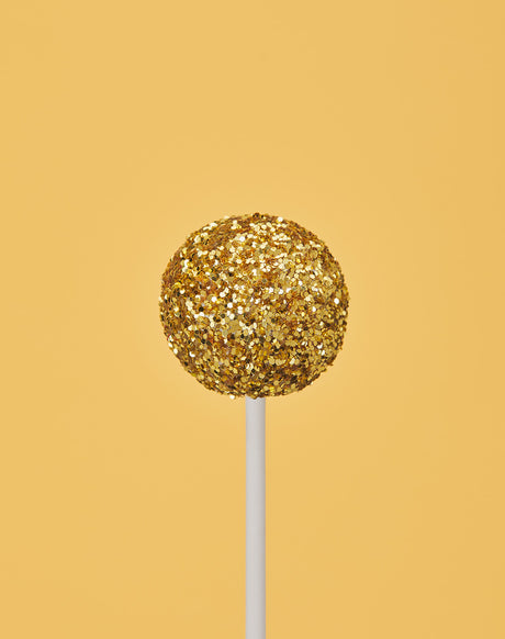 GLITTER DIET YELLOW: A single golden glitter cake pop stands on a white stick against a bright yellow background. The cake pop is round and covered in gold glitter, creating a festive and celebratory look. The simple background highlights the beauty of the cake pop and its shimmering details.