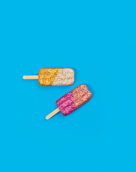 GLITTER ICE CREAM BLUE: A flat lay image showcases two popsicle treats on a vibrant blue background. The popsicles are coated in shimmering glitter, with one featuring gold and white glitter and the other sporting pink and gold. The simple composition highlights the fun and playful nature of these festive summer treats.