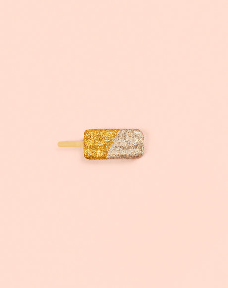 GLITTER ICE CREAM GOLD: A single popsicle coated in gold and silver glitter sits on a pink background. The popsicle is positioned horizontally, with its stick extending to the left. The background is a simple, solid pink color.