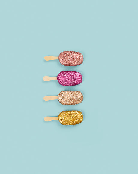 GLITTER MAGNUMS: A close up of four popsicles on a blue background. Each popsicle is covered in glitter, with the colors being pink, rose gold, gold, and a darker pink. They are arranged in a vertical line, with their sticks facing left.