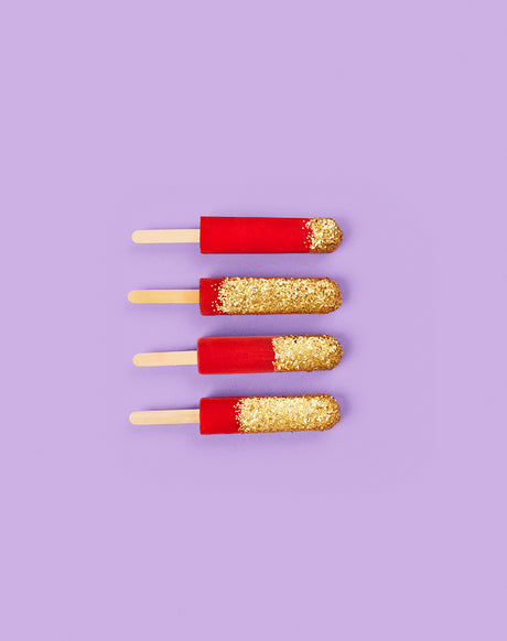 GLITTER POPSICLES 1: This image shows four popsicles arranged in a row on a purple background. The popsicles are red with gold glitter on the top half. The image is taken from a top-down view and features a simple, clean aesthetic.