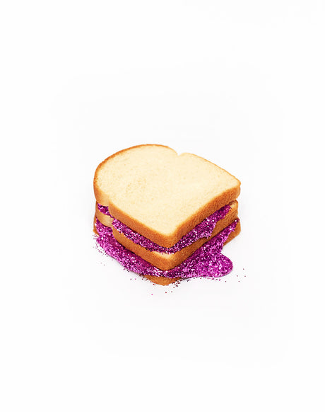 GLITTER SANDWICH WHITE: A close-up photograph of a white bread sandwich with pink glitter filling. The sandwich is sitting on a white surface, and the glitter is spilling out of the sides. The sandwich appears to be a simple, white bread, with no other visible fillings besides the glitter.