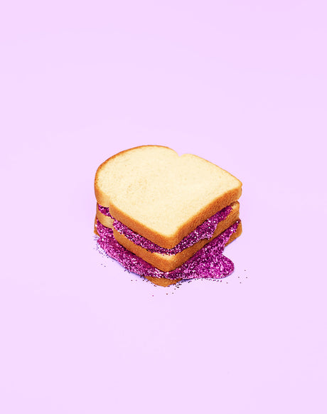 GLITTER SANDWICH PURPLE: A simple sandwich with three slices of bread and a filling made of purple glitter. The sandwich is on a lavender background and the glitter is spilling out of the sides of the sandwich.