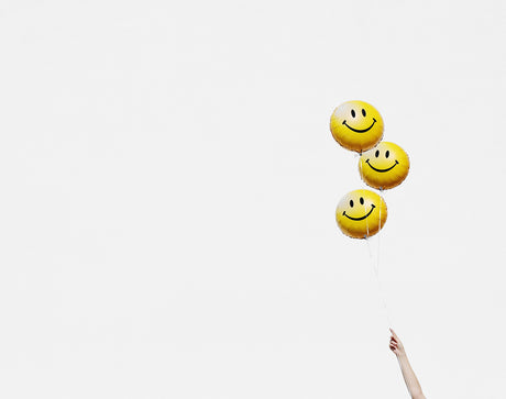 HAPPY BALLOONS: Three yellow smiley face balloons float in the air against a bright white sky. A hand is reaching up to hold the strings of the balloons.