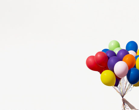 RAINBOW BALLOONS 1: A hand holds a bunch of colorful balloons against a white background. The balloons are all different colors, including red, orange, yellow, green, blue, purple, and pink. The balloons are all inflated and tied together with string.