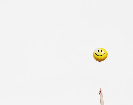 SINGLE HAPPY BALLOON: A hand reaches up towards the sky holding a yellow smiley face balloon. The balloon is inflated and has a large, black smiley face drawn on it. The sky behind the balloon is a solid white color, making for a simple yet eye-catching image.