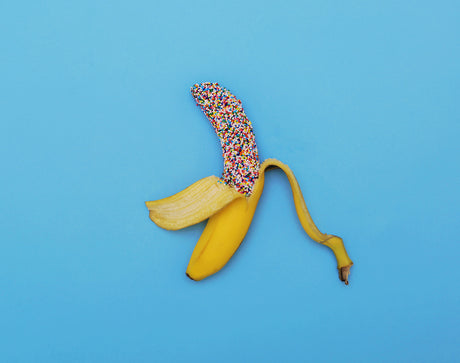 SUGAR HIGH: This image shows a single yellow banana, partially peeled, with a colorful sprinkle topping. The banana is laid horizontally on a plain light blue background. The banana is a classic, everyday food and the sprinkles add a playful, whimsical touch.