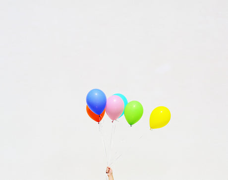 UNTITLED (BALLOONS): A photo of a hand holding a bunch of colorful balloons against a white cloudy sky. The balloons are blue, pink, green, yellow, and orange.