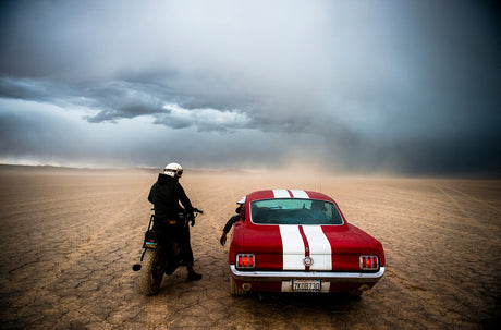 DESERT RACE: A motorcyclist in a white helmet stands next to a red classic Mustang with white racing stripes on a desert landscape. The sky is overcast and a dust storm is blowing, obscuring the distant horizon.