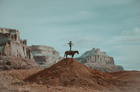 LASSO: A cowboy stands atop a desert hill, silhouetted against the vast, red rock canyons in the distance. He is holding a lasso in his right hand and gazing out towards the panorama. The horse he is riding stands calmly, its head tilted down. The sky is a clear blue, and the mountains are bathed in the warm light of the setting sun. This image evokes a sense of freedom, adventure, and the wild beauty of the American West.