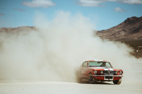 MUSTANG: A red Ford Mustang classic car is seen driving through a large dust cloud in the desert. The car is kicking up a lot of dust as it accelerates, leaving a cloud in its wake. The car is facing the camera, and the dust cloud is obscuring the background.