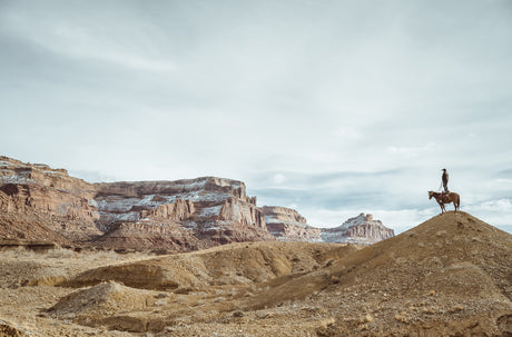 STANDING COWBOY: The image shows a cowboy on horseback standing on a hilltop in a desert landscape. The desert is characterized by red rock formations and a light dusting of snow. The sky is cloudy and there is a vast open space surrounding the cowboy.