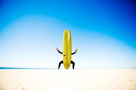 SURF FLY: A surfer stands on a white sandy beach with a yellow surfboard held straight up in front of them. Their arms are outstretched and their body is in a crouched position, as if they are about to take off on a wave. The beach is empty and the sky is a bright blue.