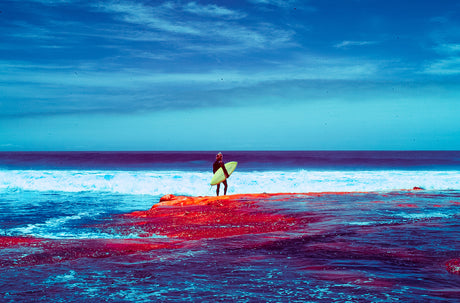 SURFING IN RED AND BLUE: A lone surfer stands on a red rock formation with a surfboard in hand, looking out at the blue ocean. The sky is a bright blue with white clouds.