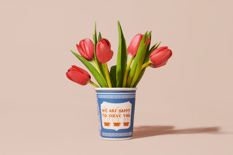 WE ARE HAPPY TULIPS: A bouquet of red tulips sits in a blue and white cup with a message printed on the front. The cup is a take on a classic diner design, featuring the phrase We Are Happy To Serve You and three stylized coffee cups with steam rising from them. The tulips are in focus, their vibrant red color contrasting with the bright blue and white of the cup. The background is a muted pale peach, allowing the flowers and cup to stand out.