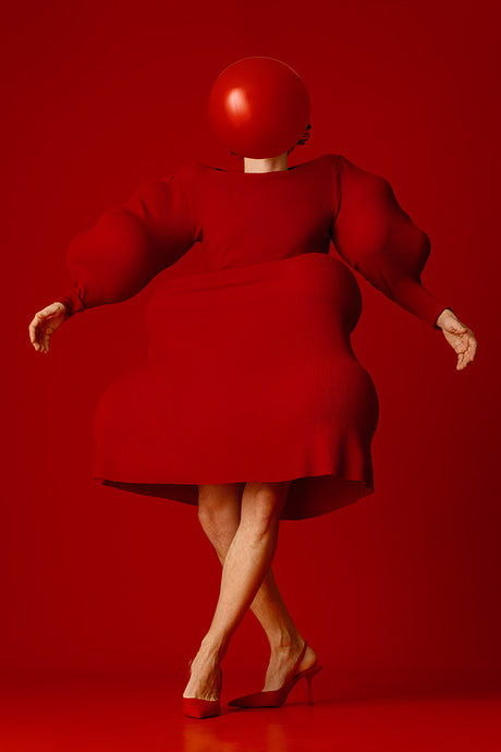 BALLOON 4: This image shows a woman in a red dress with her head covered by a red balloon. The woman is standing on a red background and her legs are crossed in front of her. She is wearing red heels and her pose is slightly dramatic, as if she is moving or about to move.