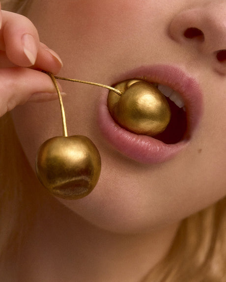 I LOVE YOU CHERRY MUCH: A close-up image of a womans mouth with two golden cherries. The woman is holding the cherries in her hand, and one is resting in her mouth. The cherries are shiny and golden, and the womans lips are pink and full. The image is suggestive of luxury and sensuality.