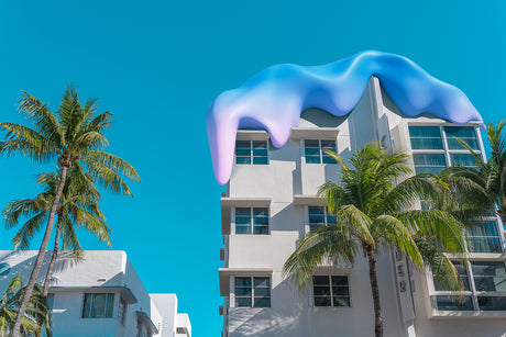 SWEET SURRENDER 2: This photograph shows an Art Deco building in Miami Beach with a unique, dripping blue roof. The building is situated on a street lined with palm trees, which create a tropical vibe. The bright blue sky creates a stunning contrast to the white building and the vibrant blue roof.