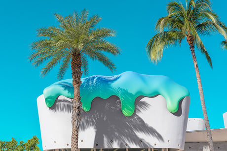 SWEET SURRENDER 3: This image shows two tall palm trees growing in front of a building in Miami with a unique design. The building features a large, white wall with dripping turquoise and green paint. The sun casts shadows from the palm trees on the building.