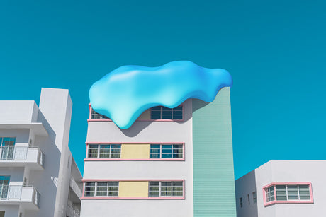 SWEET SURRENDER 4: A large blue cloud sculpture sits atop a white Art Deco building in Miami. The sculpture is made of a soft, flowing material and casts a shadow on the buildings facade. The building has a pastel pink trim and windows with a view of the clear blue sky.