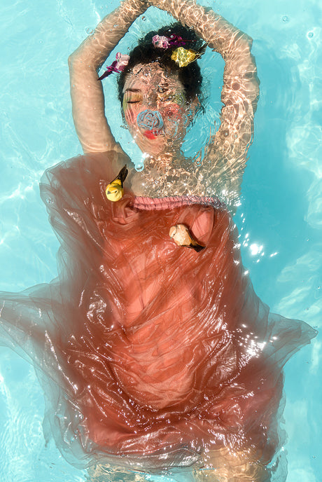 AQVA BIRDS: This image shows a woman floating in a swimming pool. She is wearing a pink dress that is partially submerged in the water. The water is clear and blue, and the womans hair is adorned with flowers.
