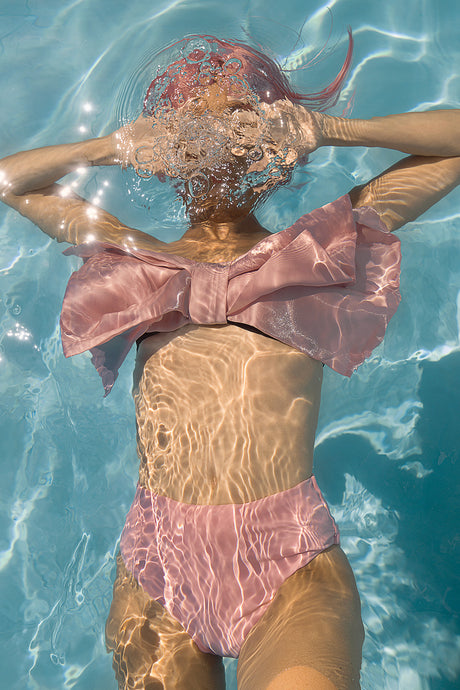 AQVA BIG BUBBLE: A woman with pink hair floats in a swimming pool, her face obscured by bubbles. She is wearing a pink swimsuit with a large bow on the top. Sunlight shines through the water, creating a shimmering effect.