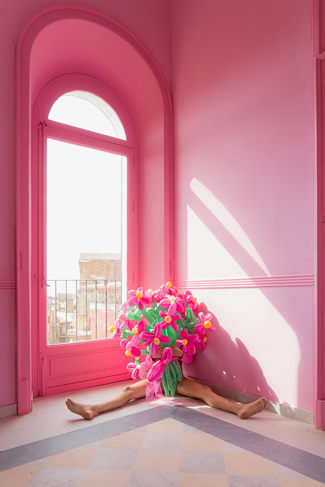 BOUQUET: A person lies in the corner of a room with pink walls and a window. They are obscured by a large bouquet of pink and green balloon flowers. The room is brightly lit, with sunlight streaming through the window.