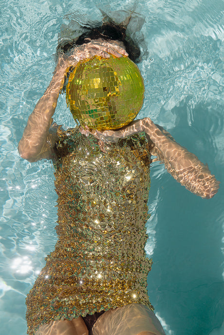 LIGHT UP GOLD: This image features a woman wearing a gold dress, submerged in a swimming pool. She is holding a large disco ball in front of her face, covering her eyes. The womans hair is dark and wet, and her body is partially visible through the clear water.