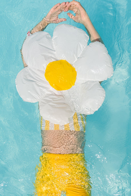 MARGARITA: A woman in a yellow swimsuit is floating on her back in a pool, relaxing with a large, white daisy pool float. The water is clear and blue, and the sunshine is reflecting off the surface. The woman appears to be enjoying a carefree summer day.